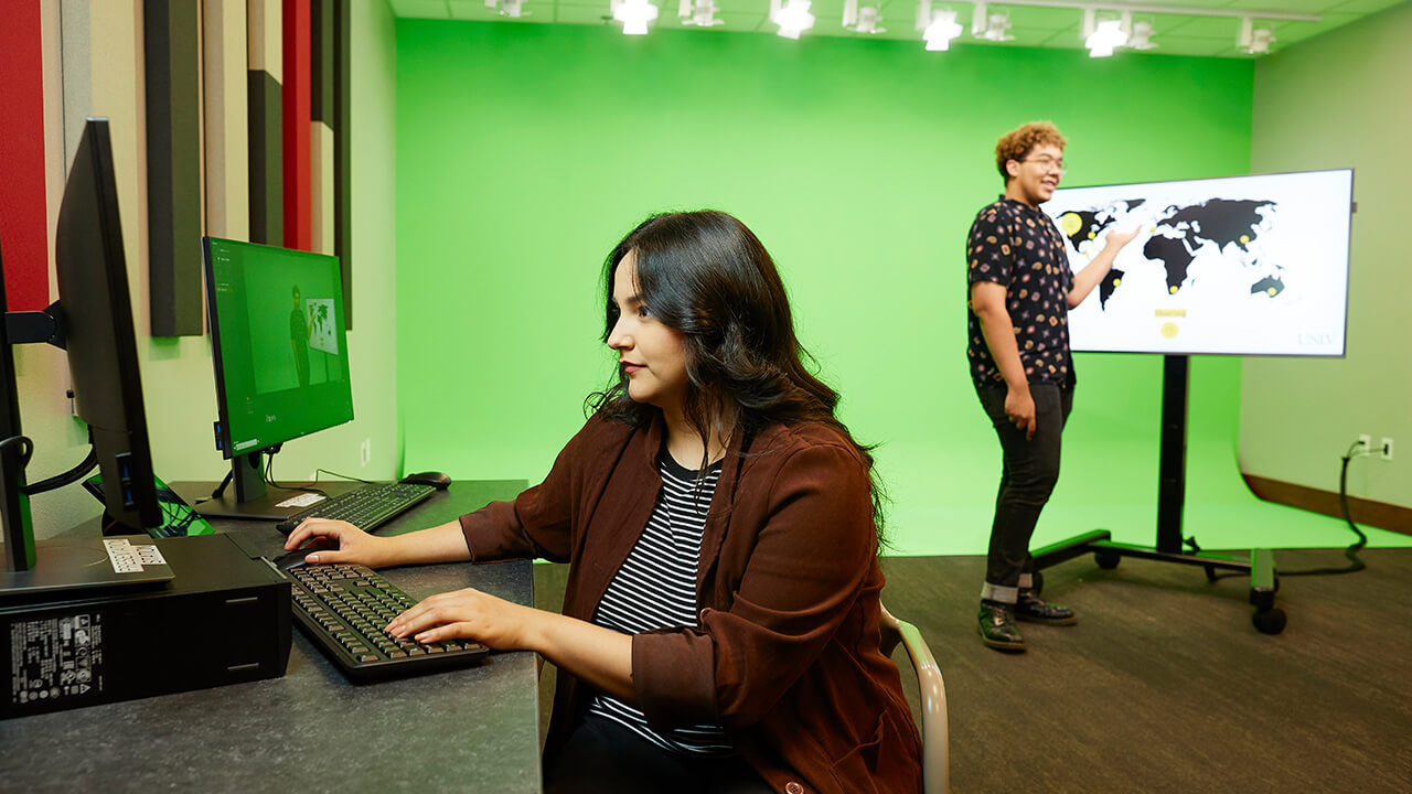 Woman sitting at computer with recording equipment with green screen behind her