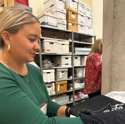 Paige Figanbaum wearing gloves, helps arrange artifacts in back stacks area with shelves of boxes in background