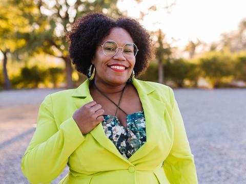 A person with curly hair wearing a lime-green blazer and glasses, smiling outdoors.