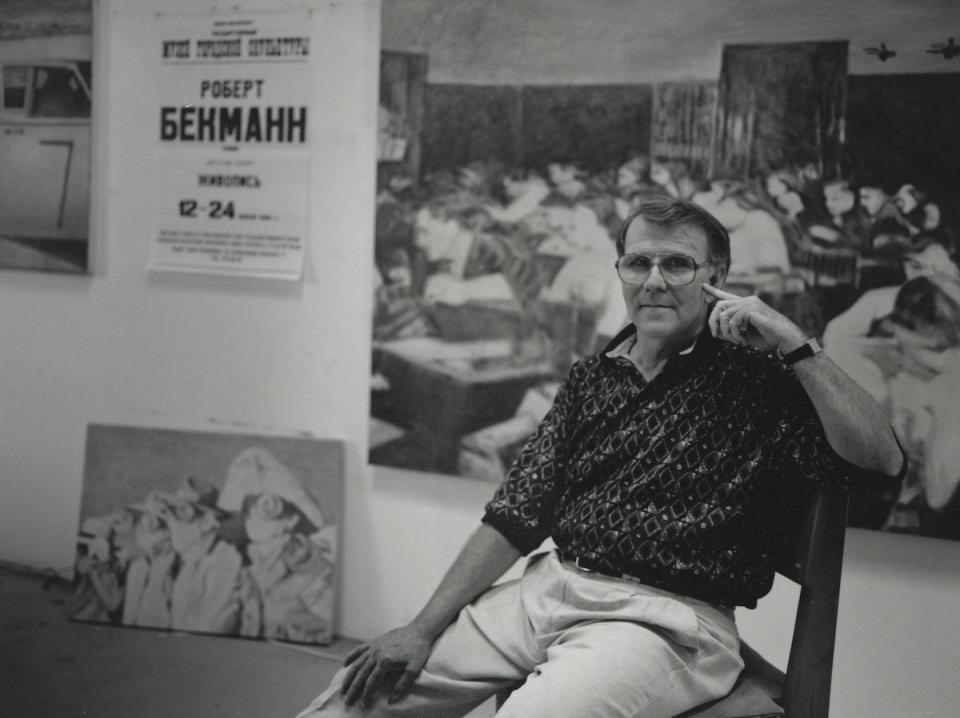 Black and white photo of Robert Beckmann sitting in front of his painted artwork.