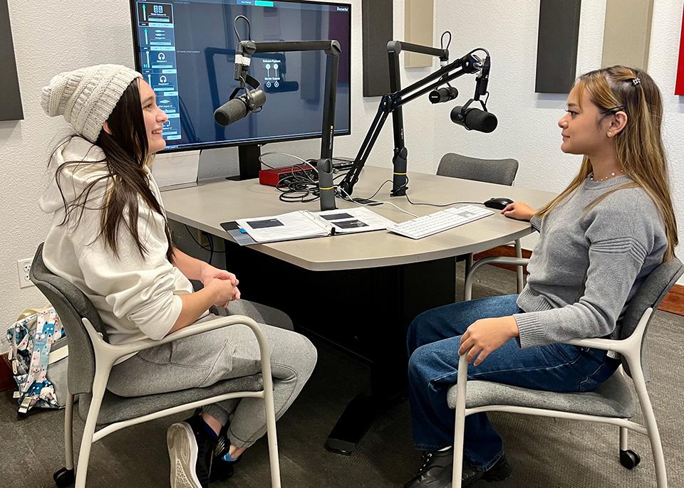 Two women converse across a desk with boom-mounted microphones in a recording studio.