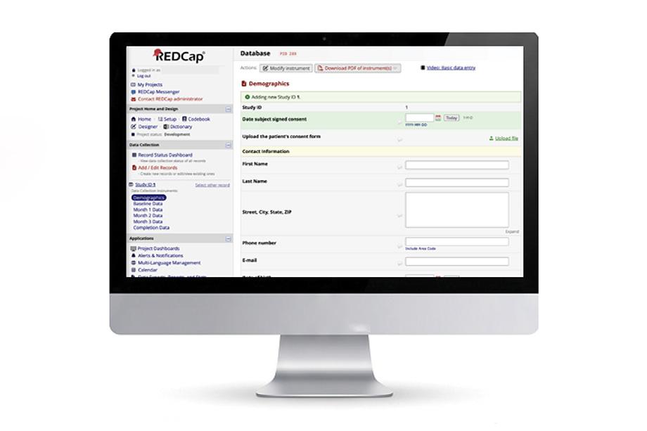 A computer monitor displaying a REDCap database interface with sections for demographics, data entry fields, and navigation options on the left.