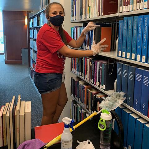 Student assistant wearing face mask and dusting book and sorting book shelves