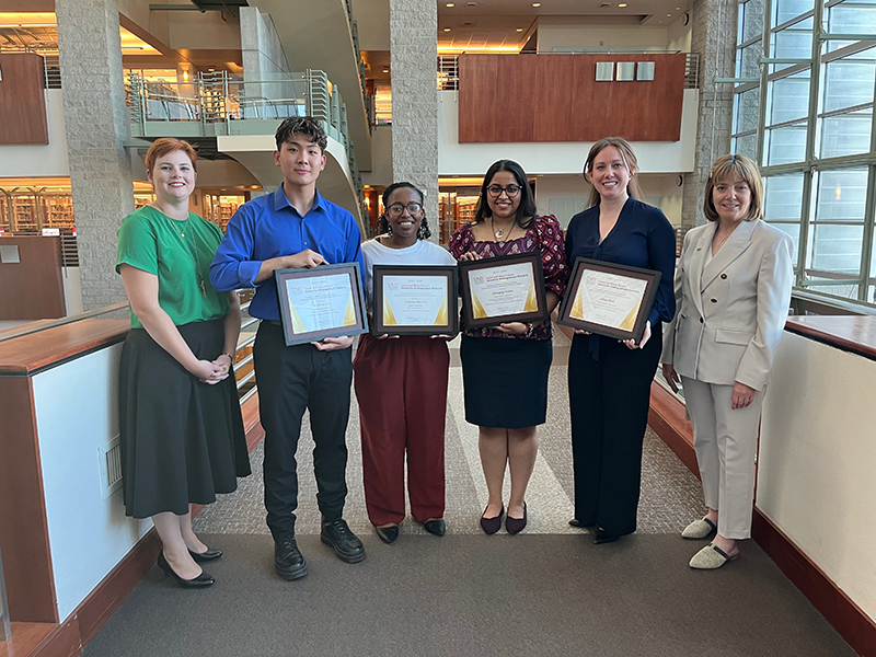 Six people standing in Lied Library, four hold framed certificates.