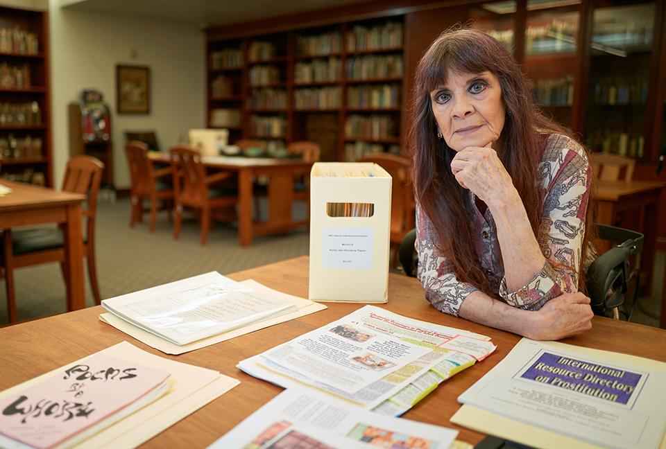 A woman sits at a table in a library with various papers and files on the table.