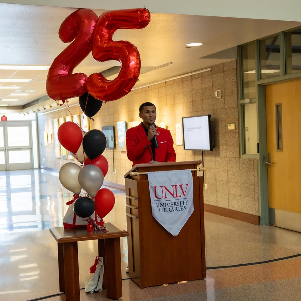 Person in a red suit speaking at a podium with "UNLV University Libraries" drape, next to red, black, and silver balloons with a large "25."