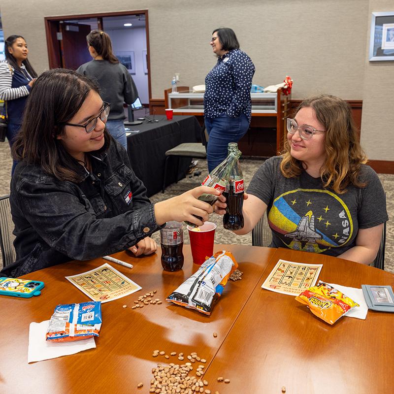 Two people clinking glass Coke bottles at a table with snacks and loteria cards.