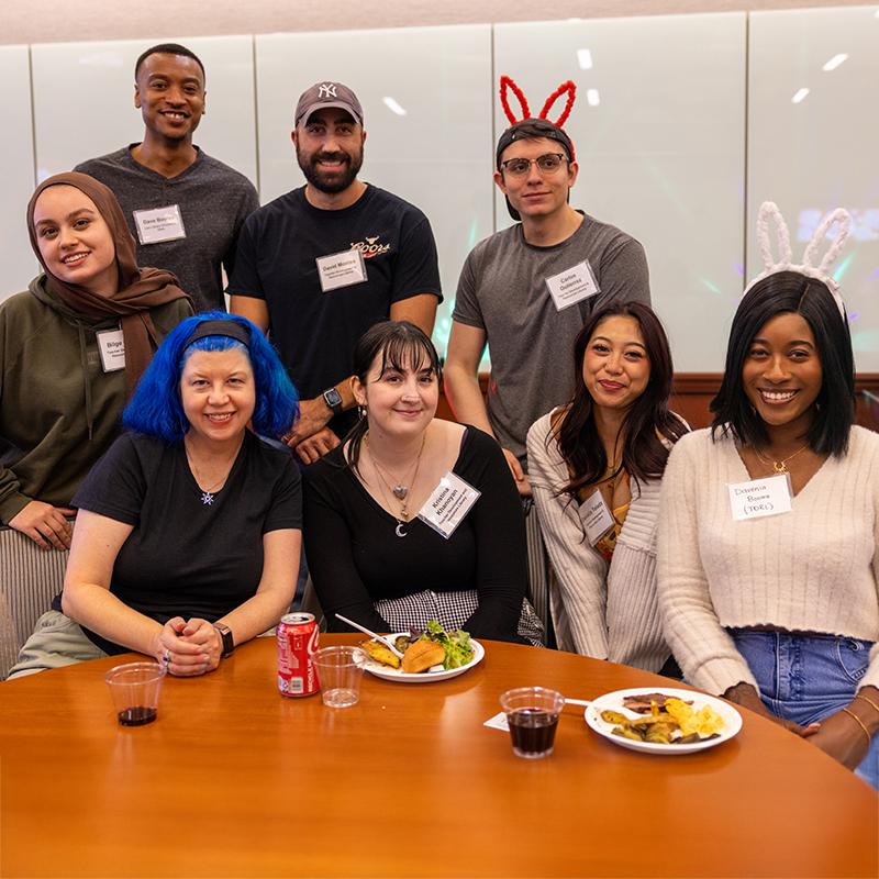 A group of eight people poses around a table with food and drinks, some wearing bunny ear headbands.