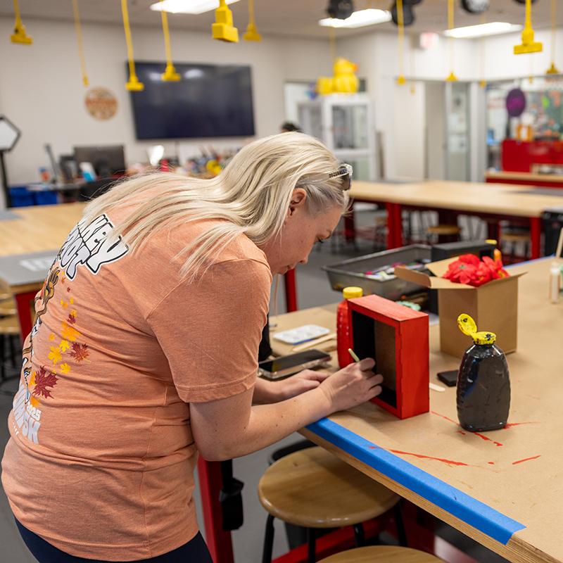 A person painting a small box red in a craft workspace with art supplies on the table.