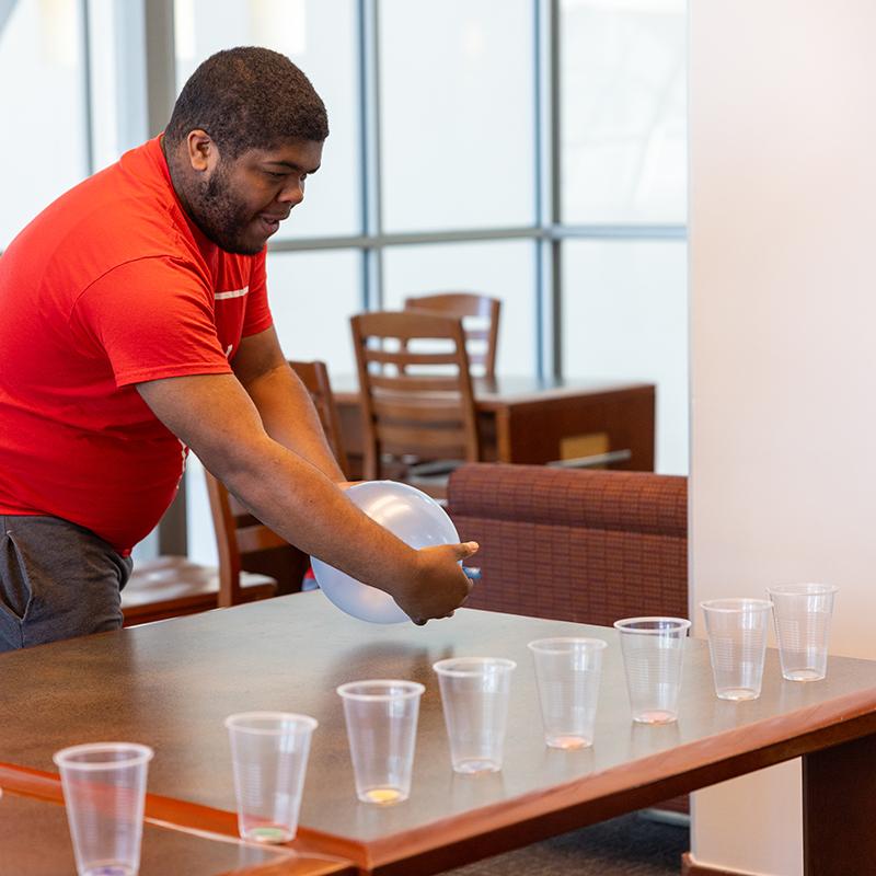 A person in a red shirt playing with a blue balloon over a table with clear plastic cups arranged in rows.