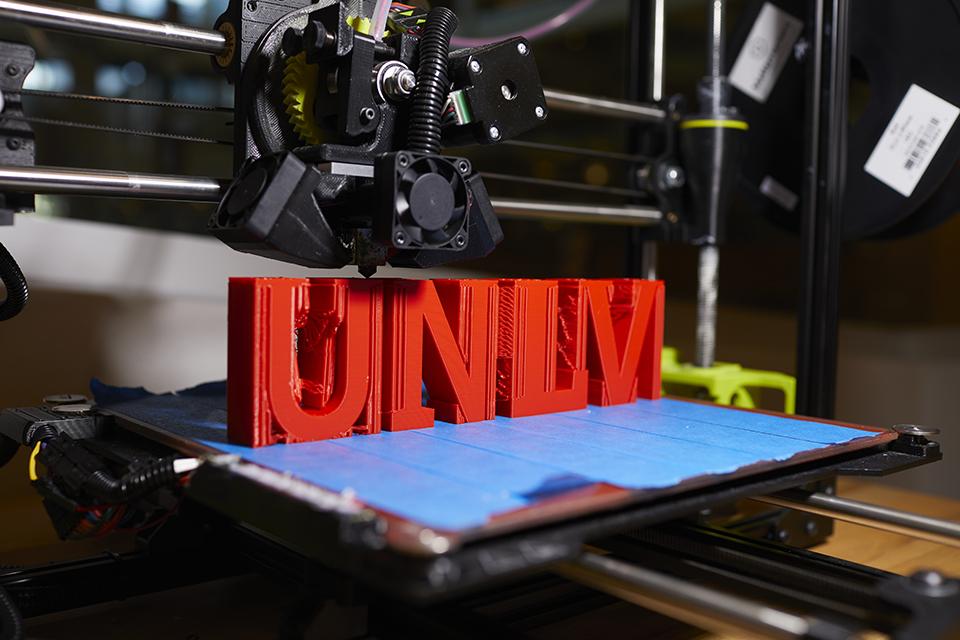 A 3D printer printing red text "UNLV" on a blue base.