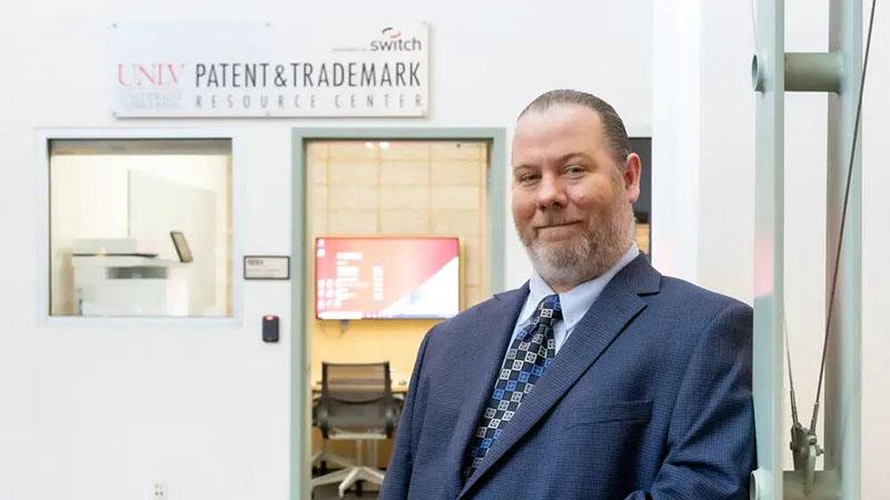 A man in a suit stands in front of the "Patent & Trademark Resource Center" with office windows in the background.