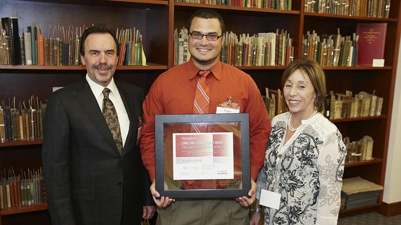 Three people in a library, the center person holding a framed certificate.