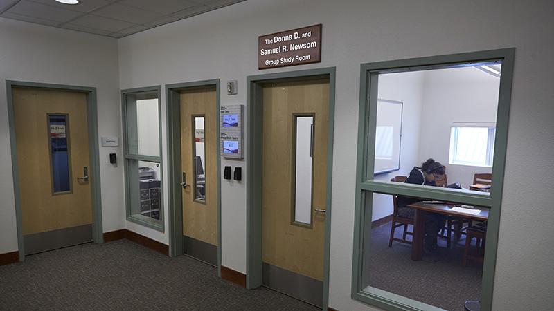 A hallway with three doors and a window showing a study room; a person is seated at a table inside.