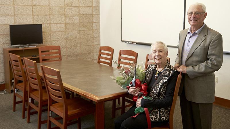 Two individuals in a study room; a woman holding flowers is seated at a table, with a man standing beside her.