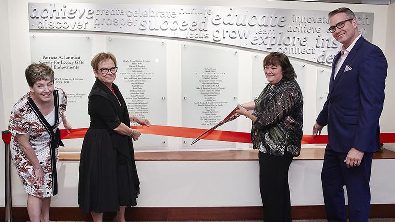 Four people participate in a ribbon-cutting ceremony in front of a display with frosted glass panels and engraved motivational words.