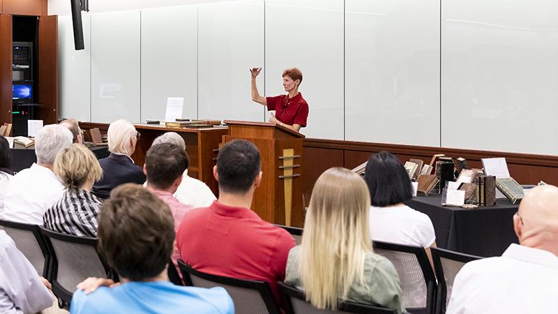 A person speaks at a podium in a lecture hall, with an audience seated and tables of books visible.