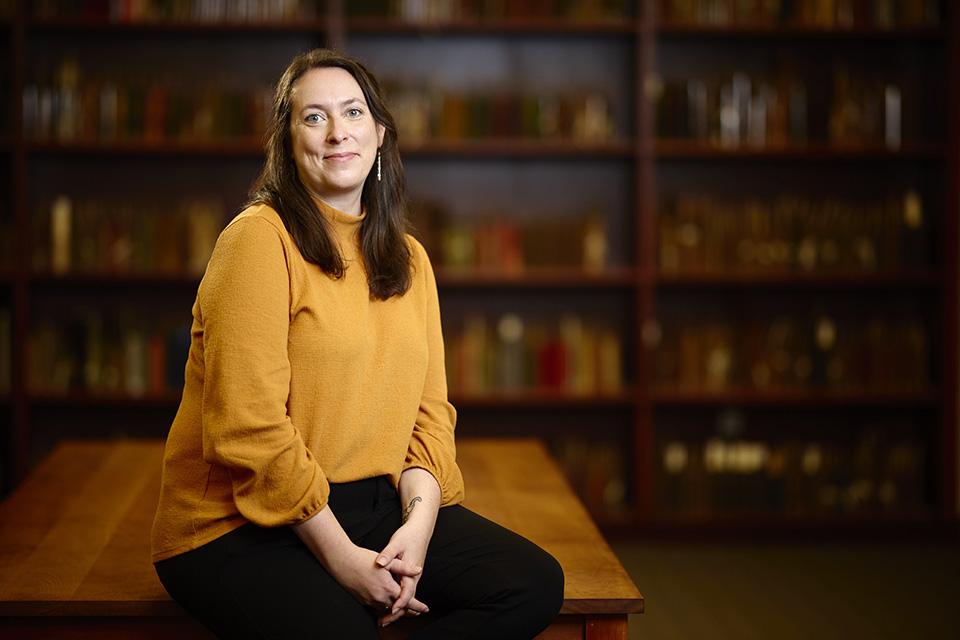 A person in a mustard-yellow sweater sits on a wooden table in front of blurred bookshelves.