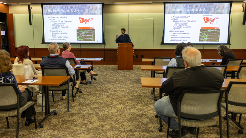 Speaker standing at a podium addressing an audience with presentation slides projected on screens.