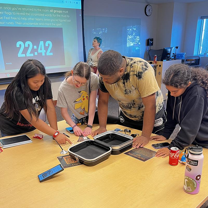Four people collaborate over a table with containers and papers; a screen shows "22:42" in the background.