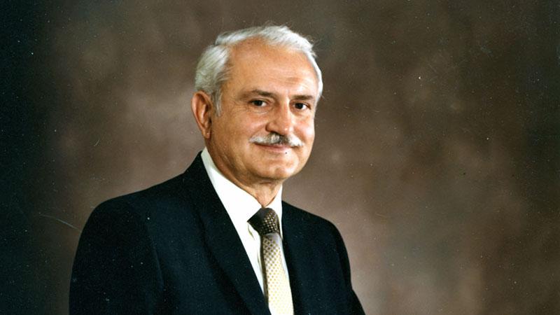 Man in a dark suit with a white mustache and white hair against a brown background.