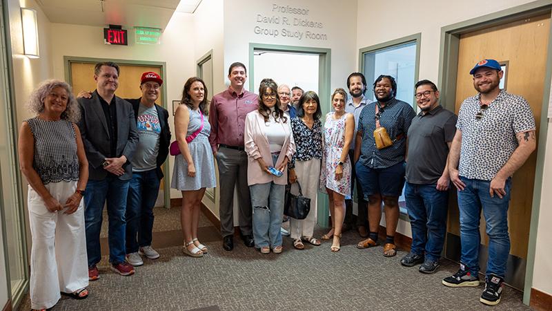 A group of fourteen people posing in a hallway near a sign that reads "Professor David R. Dickens Group Study Room."