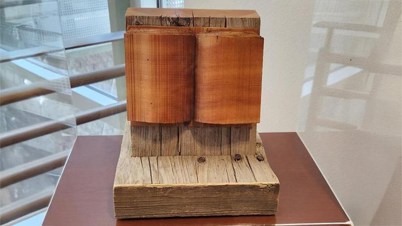 Wooden sculpture resembling an open book on a pedestal with a windowed, reflective background.
