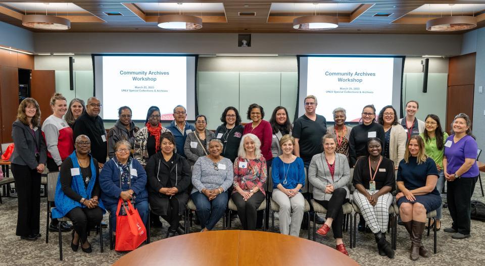 Group of about 25 workshop participants posing in a meeting room with presentation screens behind them reading “Community Archives Workshop.”