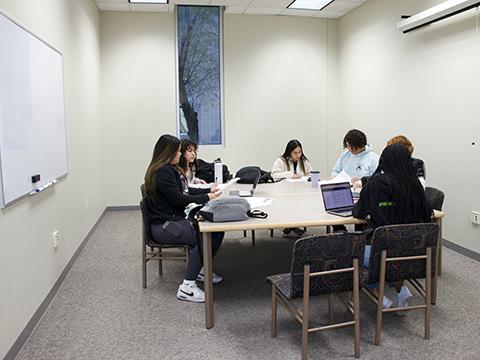 Students in study room sitting at a square table with a waiteboard, pull down screen, and vertical window.