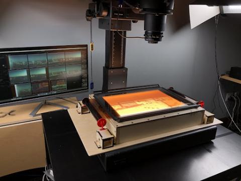 Placed on the table top of our Phase One Rapid Capture System, the negative carrier is composed of a wooden base, placed directly underneath a camera where two pieces of anti-newton ring glass hold the negative in place. An orange-tinted negative of the Las Vegas Strip is stretched across its surface. Beneath the base, a light box is turned on illuminating the negative.