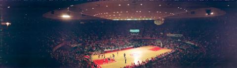 A college basketball game between UNLV and Kentucky inside the Convention Center in Las Vegas, Nevada. The image is taken from high up in the stand behind a packed crowd watching the game.