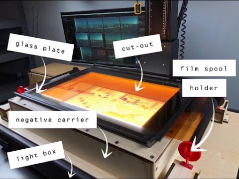 n up close view of a negative carrier with a negative of the Las Vegas Strip stretched out across its surface. The negative is illuminated by a light box placed underneath the negative carrier. A computer monitor in the background shows the digitized images of the negative.