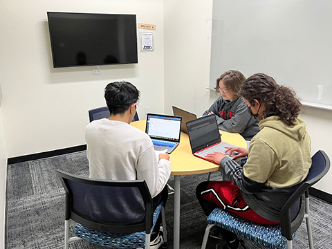 Three students sitting at a round table with a laptops. Room has a wall-mounted TV monitor and glassboard.