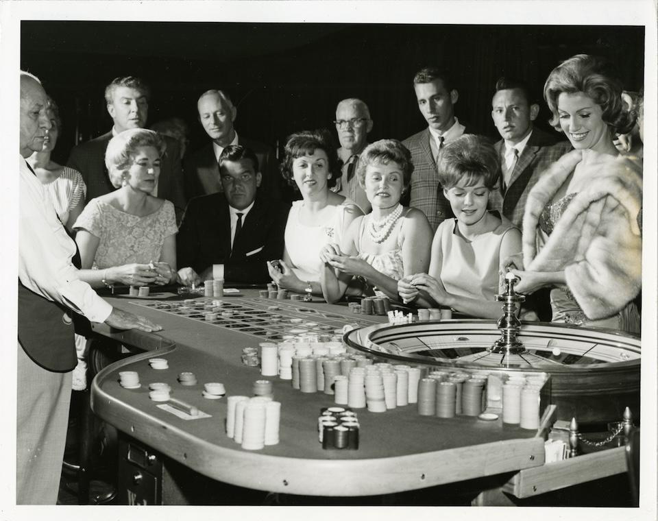 A group of men and women around a casino roulette table at the Sands Hotel, with chips spread across the surface.