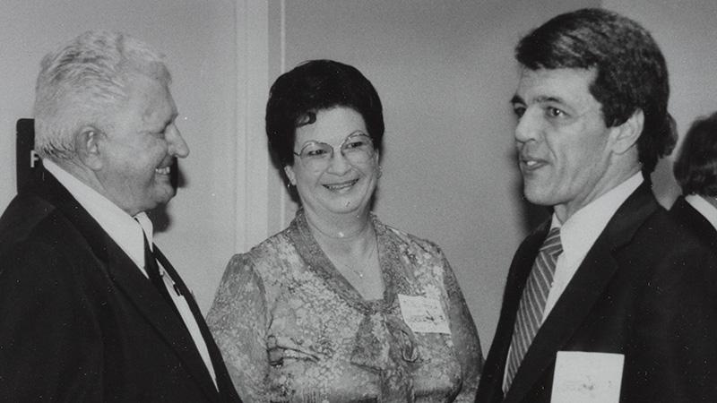 Three people conversing at an event, wearing formal attire and name tags.
