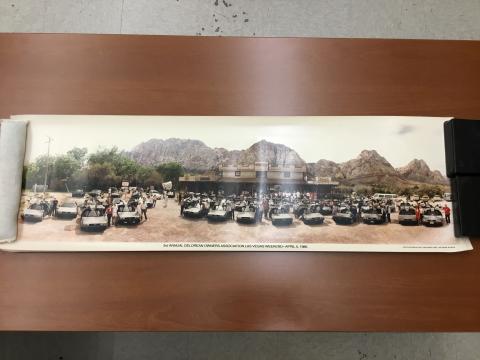 A large gathering of Delorean cars parked in front of Bonnie Springs near Las Vegas, Nevada. The cars are organized into several rows and face the camera. Owners pose beside their cars for the picture. Red Rock Canyon can be seen in the background