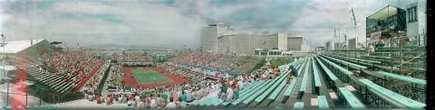 A distant view of an outdoor tennis match between Johan Kriek and Jimmy Arias at the Caesars Palace in Las Vegas, Nevada. A crowd of people sitting in the stands encircles the match. The Caesars Palace is in the background.