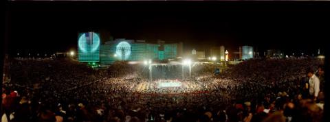  A distant view of a boxing match between Larry Holmes and Muhammad Ali outside of the Caesars Palace in Las Vegas, Nevada. A large crowd circles and watches the match. The exterior of Caesars Palace is illuminated in the background.)