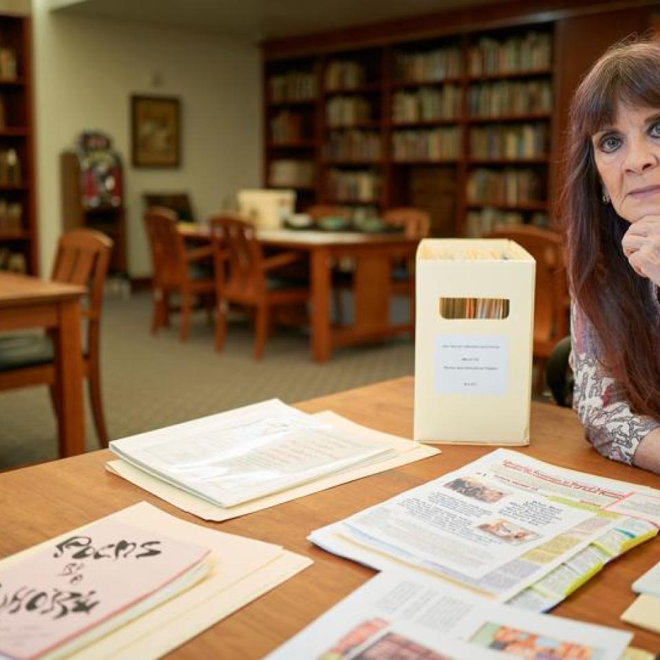 A woman sits at a table in a library with documents spread out in front of her.