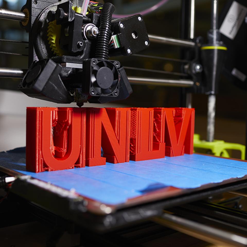 A 3D printer printing red text "UNLV" on a blue base.