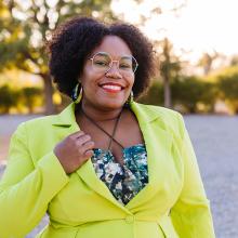 A person with curly hair wearing a lime-green blazer and glasses, smiling outdoors.