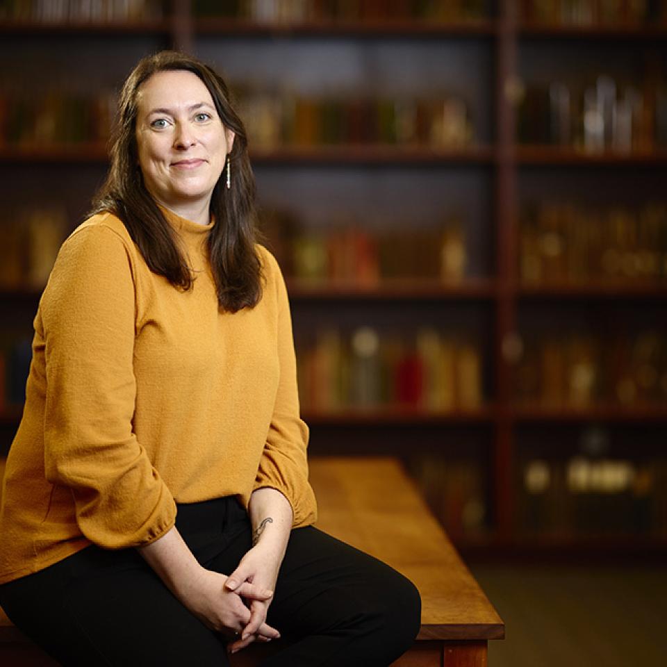 A person in a mustard-yellow sweater sits on a wooden table in front of blurred bookshelves.