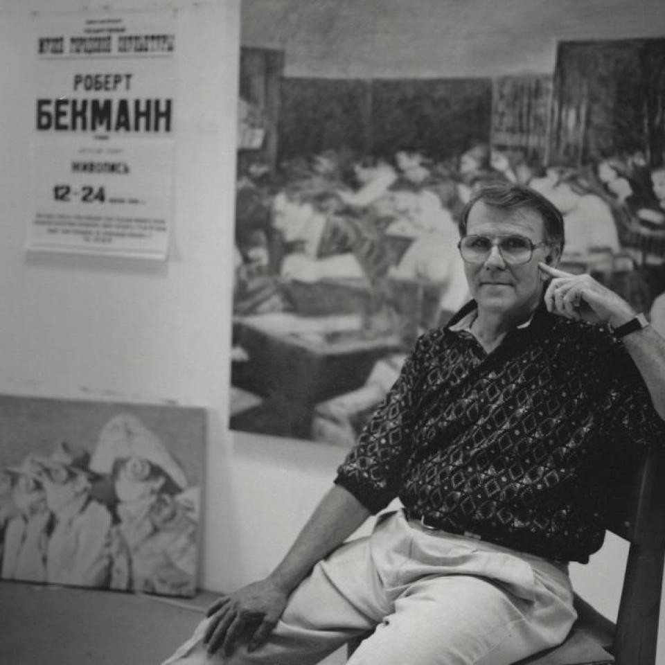 Black and white photo of Robert Beckmann sitting in front of his painted artwork.