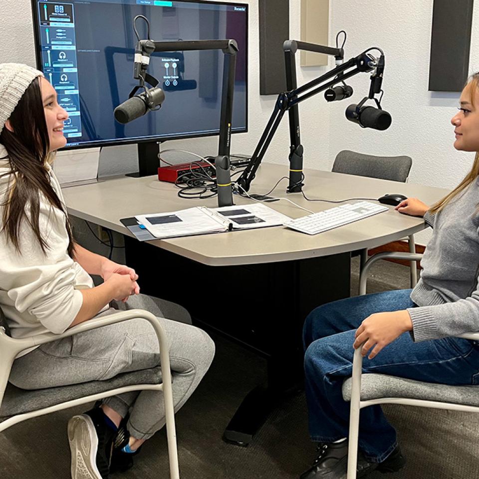 Two women converse across a desk with boom-mounted microphones in a recording studio.
