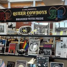 Library display titled "Queer Cowboys: Branded with Pride" featuring cowboy memorabilia and rainbow flag.